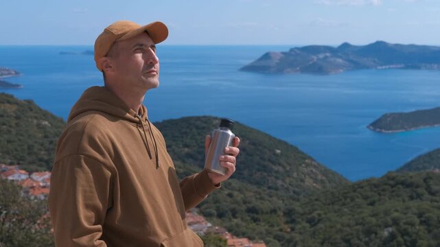 Hiker holding reusable water bottle enjoying sea view. Young man in a hoodie and cap enjoying the stunning sea view from a mountain top, holding a reusable metal water bottle and basking in the sun