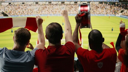 Soccer supporters in red jerseys watch the field from stadium stands from behind, waving scarves, chanting on the terrace and celebrating active team spirit during a live football match. © DC Studio