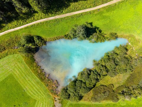 Aerial view of a vibrant turquoise pond surrounded by lush green meadows and dense trees in Sulden, Trentino-Alto Adige, Italy.