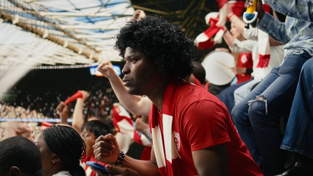 Black football fan wearing a wig monitors live betting odds at a stadium during a soccer match surrounded by cheering crowds while using a mobile sportsbook app with anticipation and excitement.