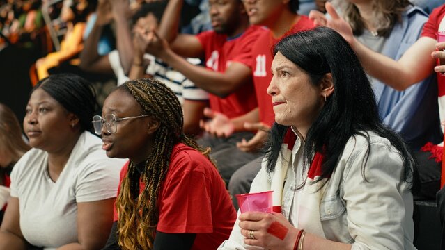 Worried mature female football supporter holds a pink cup and red scarf in stadium stands biting her lip during a tense live soccer match that highlights inclusive sports culture among diverse fans.