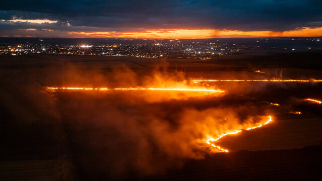 Aerial view of a field fire burning at night with bright orange flames and smoke, set against a dark landscape with distant city lights in Sremska Mitrovica, Vojvodina, Serbia.