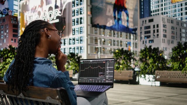 Woman crosschecks forex financial data on a laptop from a city bench near a financial district analyzing stock market trends and trade data as a remote employee working outdoors in an urban landscape.