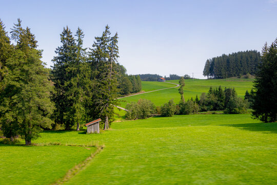 Bavarian countryside landscape, A traditional wooden barns standing alone on a rolling hill, Lush green grass meadow against majestic Alpine mountains under blue sky and white clouds, Bavaria, Germany