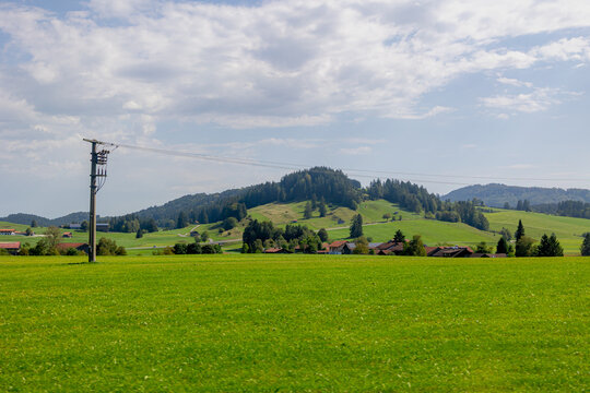 Rural Bavarian countryside landscape, Traditional wooden houses and barn on rolling hills, Lush green grass meadow against majestic Alpine mountains under blue sky and white clouds in Bavaria, Germany