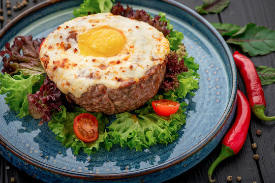 Fried egg and meat patty with tomato garnish. Green and purple lettuce frames the portion, with chili peppers beside the ceramic plate.