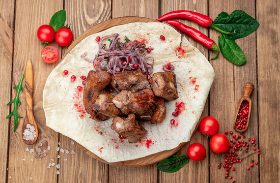 Grilled meat chunks with onion on flatbread. Pomegranate seeds garnish the portions on a round board surrounded by tomatoes and chili peppers.