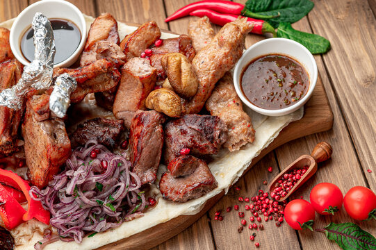 Assorted grilled meat with potatoes and onion. Chicken wings and dipping sauces frame the portions, with tomatoes beside the serving board.