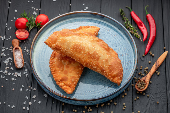 Golden fried chebureks served on blue ceramic plate. Close view of deep cooked turnovers placed on round dish surrounded by red peppers salt and green herbs.