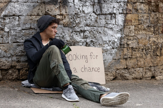 Homelessness and poverty crisis, young man sitting on street with cardboard sign looking for a chance and holding a cup for help in urban environment
