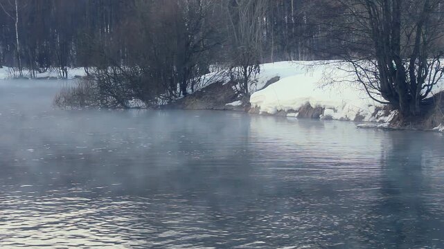 Steam rising over cold river winter forest. Snowy banks bare trees frosty mist.
