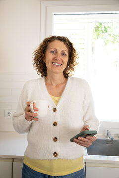 Smiling woman holding coffee and mobile phone in kitchen