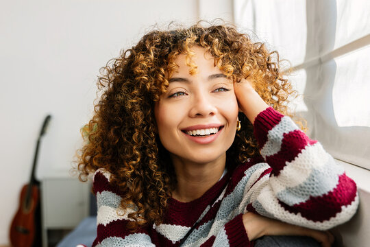 Happy young woman with curly hair relaxing on sofa at cozy home. Colombian Latin American girl enjoying winter free time indoors. People, domestic lifestyle and beauty concept.