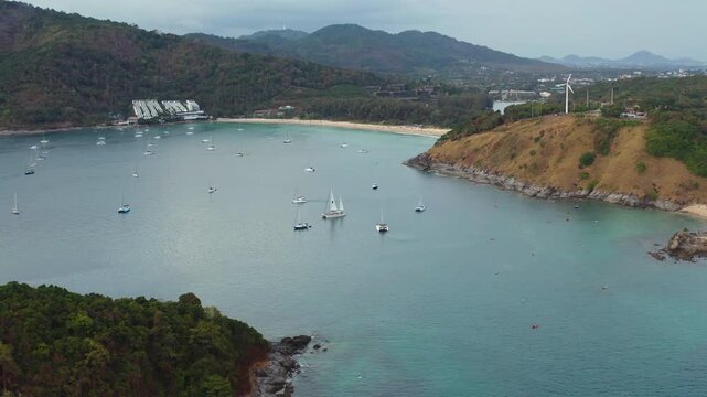 Nai Harn Beach bay in Phuket, Thailand. Aerial drone flight panorama showing numerous sailboats anchored in the turquoise waters near the sandy shore. Surrounded by lush green hills under a cloudy sky