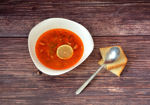 Traditional Russian solyanka soup with a lemon wedge on a wooden table, two pieces of bread and a tablespoon nearby.