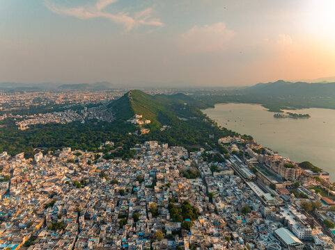 Aerial view of the historic city of Udaipur with dense urban rooftops, Lake Pichola, and surrounding green hills under the warm glow of the golden hour in Udaipur, Rajasthan, India.