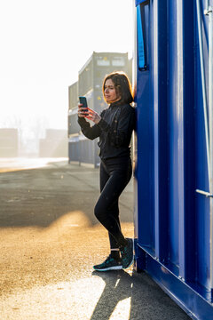 Woman using smartphone leaning on a blue shipping container in an industrial area