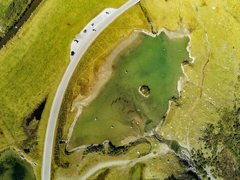 Aerial view of Silvretta-Hochalpenstrasse winding past a small alpine lake with a tiny island, surrounded by green meadows and rocky terrain in Partenen, Vorarlberg, Austria.