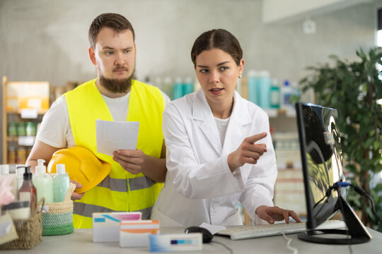 At cash register area with PC, girl pharmacist and builder visitor to pharmacy communicate. Man with doctors prescription asks to collect all the necessary drugs for course of treatment, ask questions