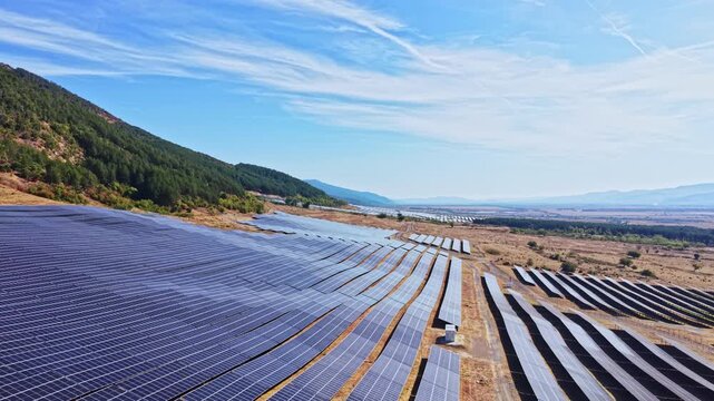 Solar panels cover a large area in a rural region. The field is flat and has rows of panels aligned in an organized manner. Hills rise in the background under a clear blue sky.