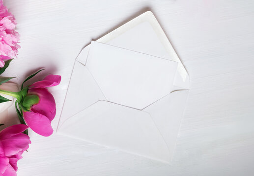 Blank card in open white envelope with pink peonies on wooden table