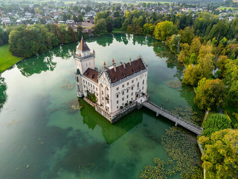 Aerial view of Schloss Anif, a historic water castle surrounded by a lush green lake, dense trees, and a wooden bridge in Anif, Salzburg, Austria.