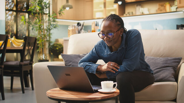 African American woman sings aloud while handling remote tasks at home, enjoying carefree telework, multitasking and a relaxed freelancer lifestyle in a domestic setting with laptop technology.