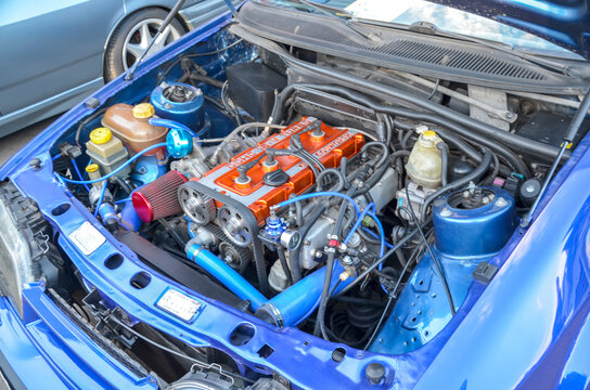 Engine bay of blue Ford Sierra RS Cosworth with bright orange DOHC 16V Turbo valve cover, exposed timing gears, blue silicone hoses, and performance tuning upgrades