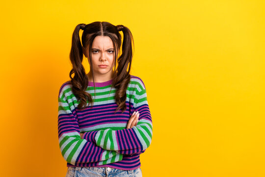 Young woman with colorful striped sweater stands against yellow background with crossed arms and a stern expression