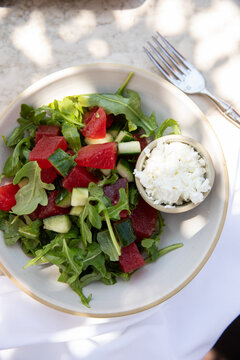 overhead shot of arugula salad with chunks of watermelon and side of feta cheese in an upscale restaurant patio