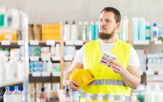 Young man builder in uniform chooses box of paracetamol tablets in pharmacy