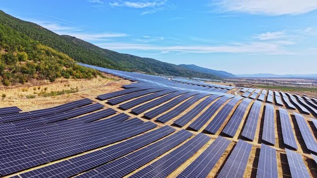 Rows of solar panels are placed on a hilly area under a blue sky with some clouds. The landscape shows a mix of hills and open land where the panels are installed.