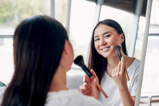 Woman smiling while applying powder with a makeup brush in front of a mirror during a relaxed morning beauty and skincare routine at home