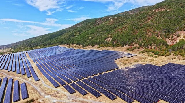 Solar panels cover a large area of land near a hillside. The installation is set against a backdrop of trees and hills under a blue sky. Clean energy generation is the focus.