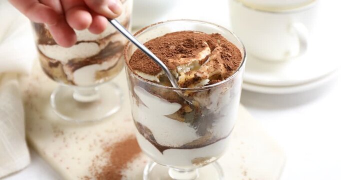 Woman eating tasty tiramisu from dessert coupe at white table, closeup