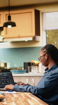 Vertical aspect: Black woman programs on laptop in kitchen during hybrid IT work, writing source code with command-line tools and cloud computing for software development.