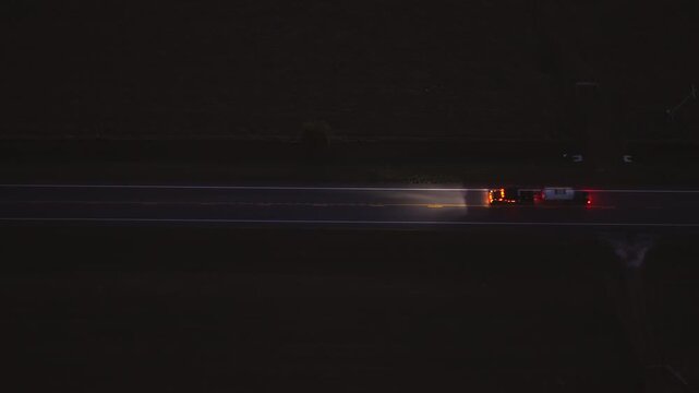 High angle view of vehicles driving on country road at twilight, illuminated by headlamps, capturing motion, travel and peaceful rural scenery.