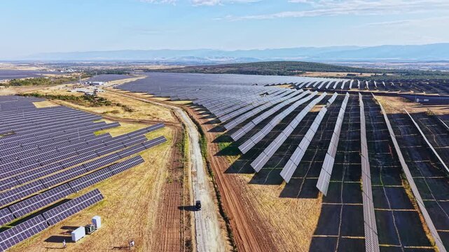 Rows of solar panels stretch across the landscape, capturing sunlight. The scene takes place in a rural area during daylight. The terrain is dry with some hills in the distance.