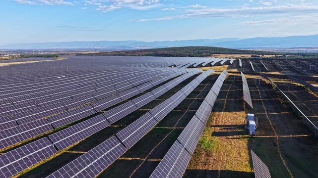 A large solar panel farm located in a rural area. Many rows of solar panels are aligned in the field. The sky is clear with some clouds, and mountains are visible in the distance.