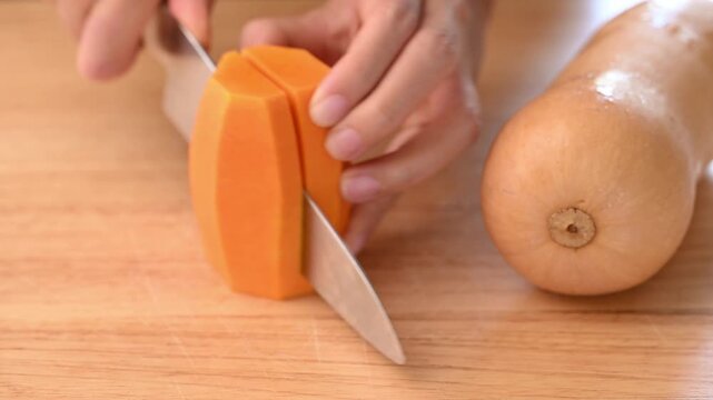 Woman hands using a kitchen knife to cut butternut squash into small pieces on a wooden board for soup preparation