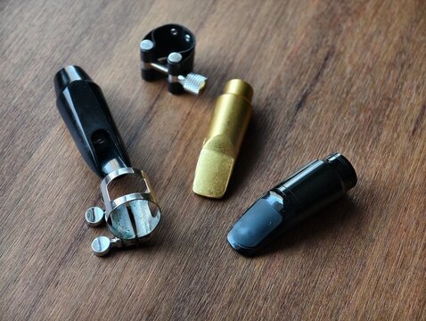 Perspective view of various woodwind instrument mouthpieces and metal ligatures placed on a natural wooden surface. Ideal for music shop catalogs, jazz music blogs, and equipment guides.