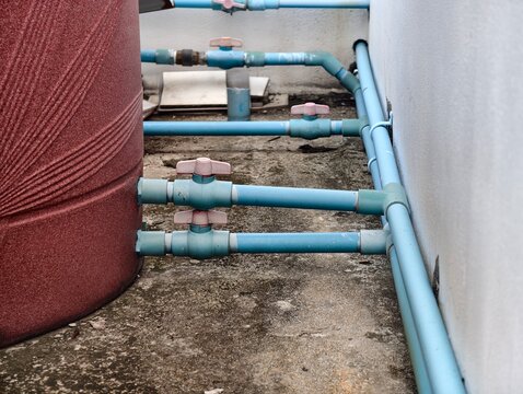A close-up shot of two blue PVC ball valves with white handles installed on water pipes, connected to the side of a large reddish-brown domestic water storage tank.