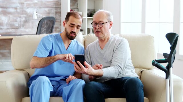 male nurse guides an older man through a smartphone while they sit together, illustrating digital assistance, communication and supportive senior care.