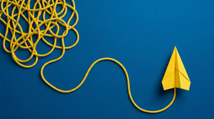 Fototapeta premium Conceptual top view of a yellow paper plane flying out from a tangled messy rope on a blue background, symbolizing finding solutions, business strategy, creative thinking, and mental clarity.