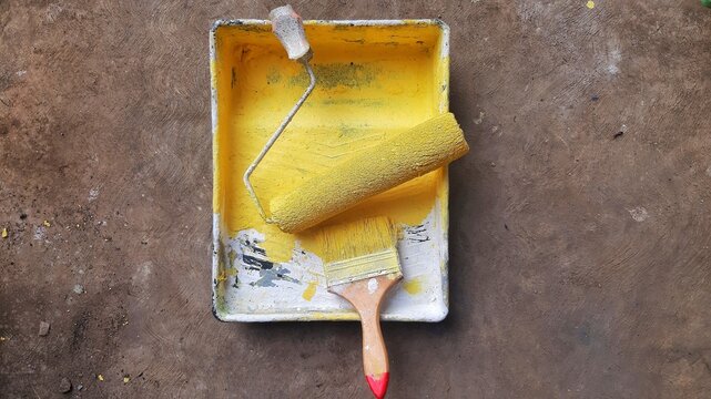 Yellow paint roller and paintbrush in a messy tray on a concrete floor during home renovation work.