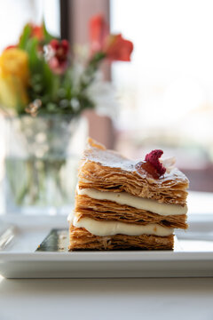 close up image of layers of mille-feuille pastry on white dish in minimal modern cafe with flowers and window in the background