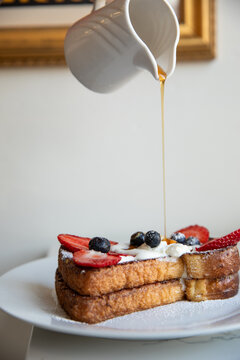 hand pouring maple syrup from white ceramic jar onto french toast with fresh berries and cream on a white plate in modern minimal cafe