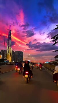 Motorcyclists riding across a city bridge at dusk with the Landmark 81 skyscraper illuminated against a vibrant pink and purple sunset sky