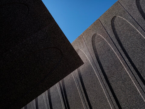 Low-angle close-up of a contemporary public building exterior, showcasing deeply textured dark granite or concrete panels with carved elongated arch motifs. 