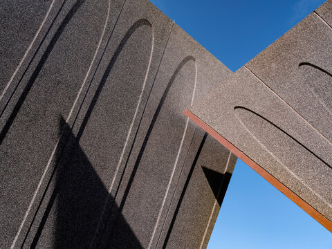 Low-angle close-up of a contemporary public building exterior, showcasing deeply textured dark granite or concrete panels with carved elongated arch motifs. 